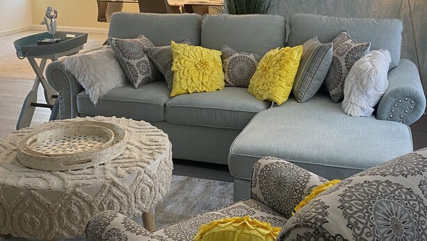 Ein Wohnzimmer mit einem grauen Ecksofa, gemusterten Sessel und einem runden Couchtisch. Im Hintergrund ist eine Essecke mit Tisch und Stühlen zu sehen.