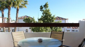 Un patio al aire libre con una mesa redonda de vidrio y sillas de metal, rodeado de paredes blancas y palmeras.