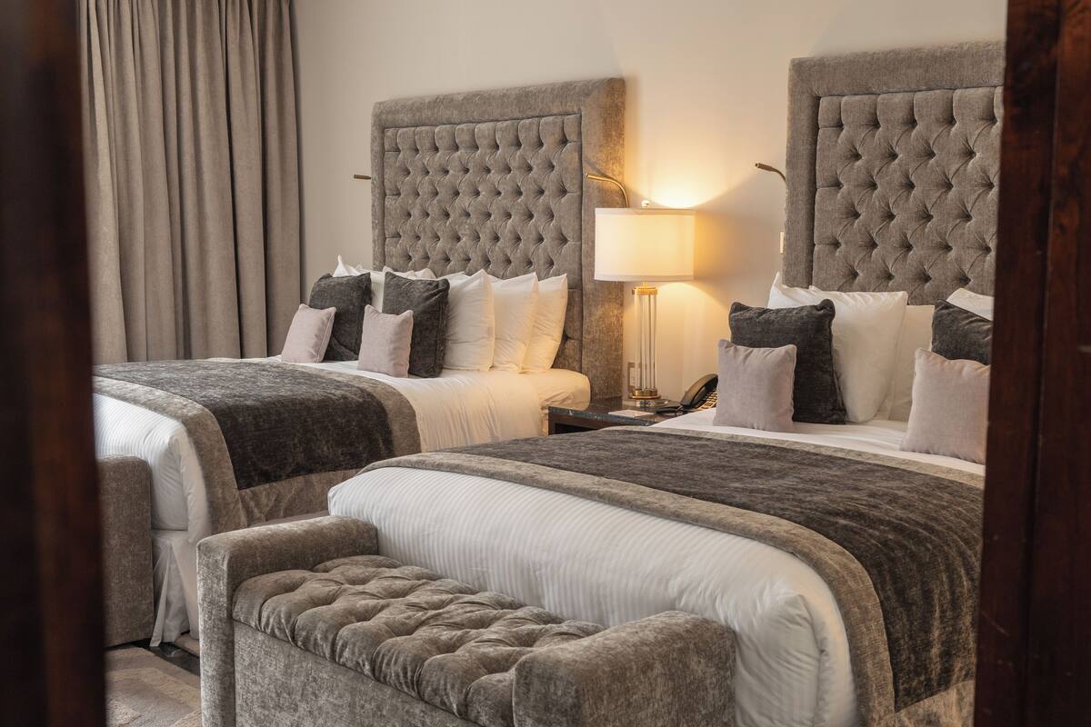 A hotel room with two beds, each with a tufted headboard, a bedside table with a lamp, and a bench between the beds.