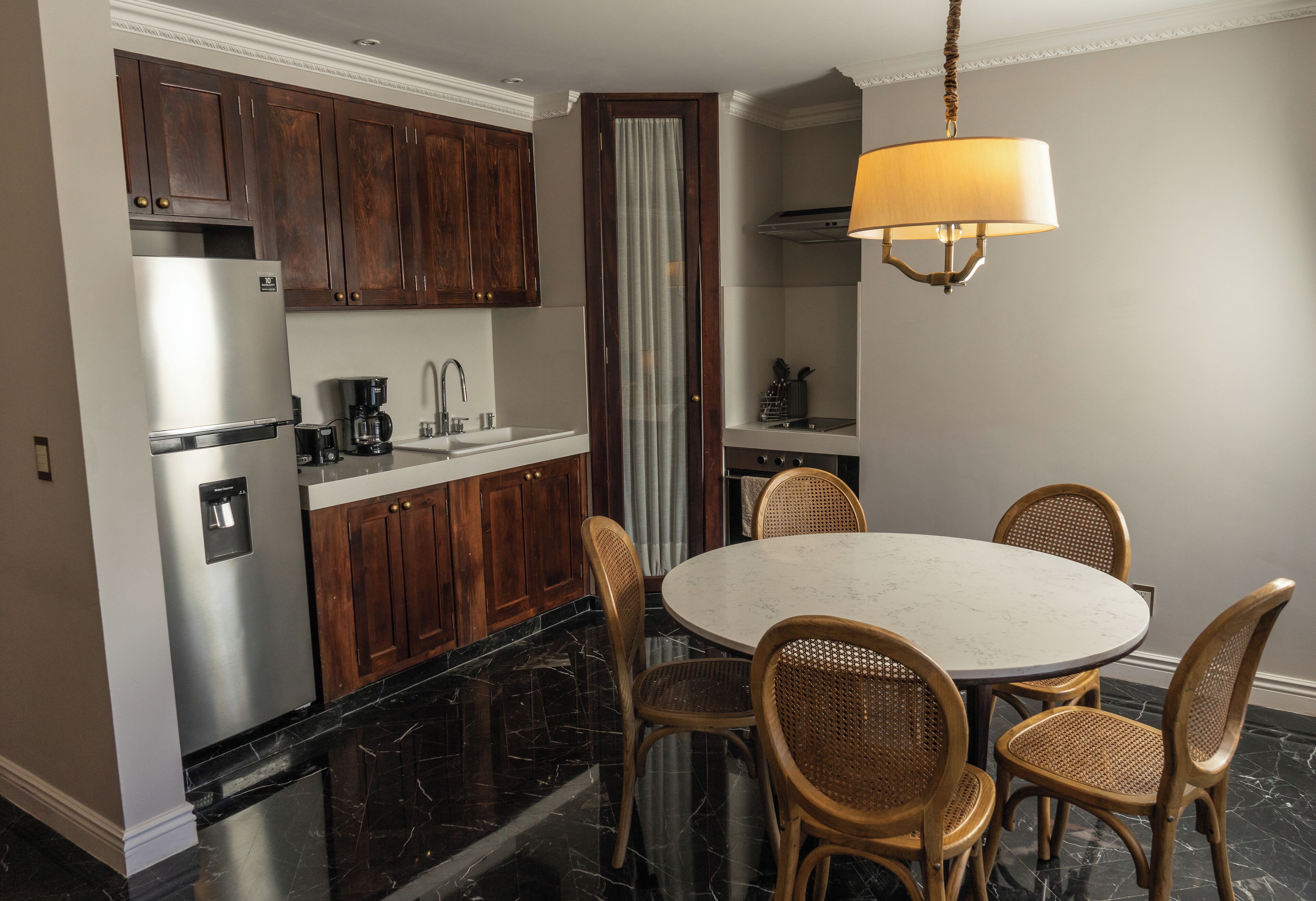 Une cuisine moderne avec des meubles de rangement en bois foncé, un réfrigérateur en acier inoxydable, une table de salle à manger ronde entourée de chaises en osier et un luminaire suspendu.
