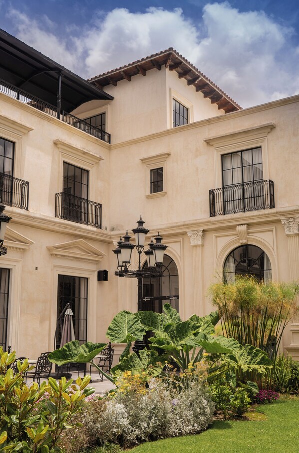 Un edificio de dos plantas con balcones, ventanas amplias y un jardín con plantas diversas y mobiliario exterior.
