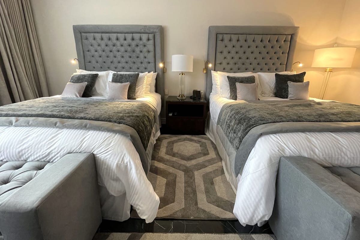 A double room with two beds, each with a tufted headboard, a bedside table with a lamp, and a patterned rug.