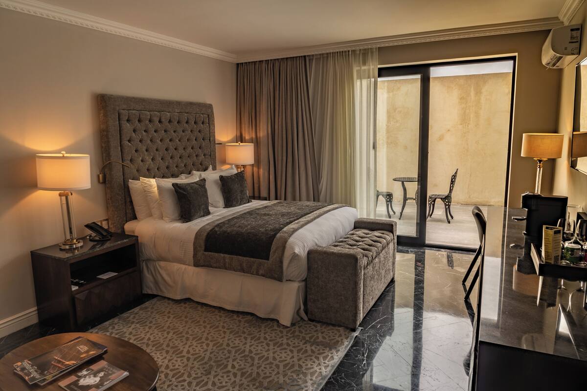 A hotel room with a large bed, two bedside tables with lamps, a small round table with magazines, a sofa, and a balcony with a chair.