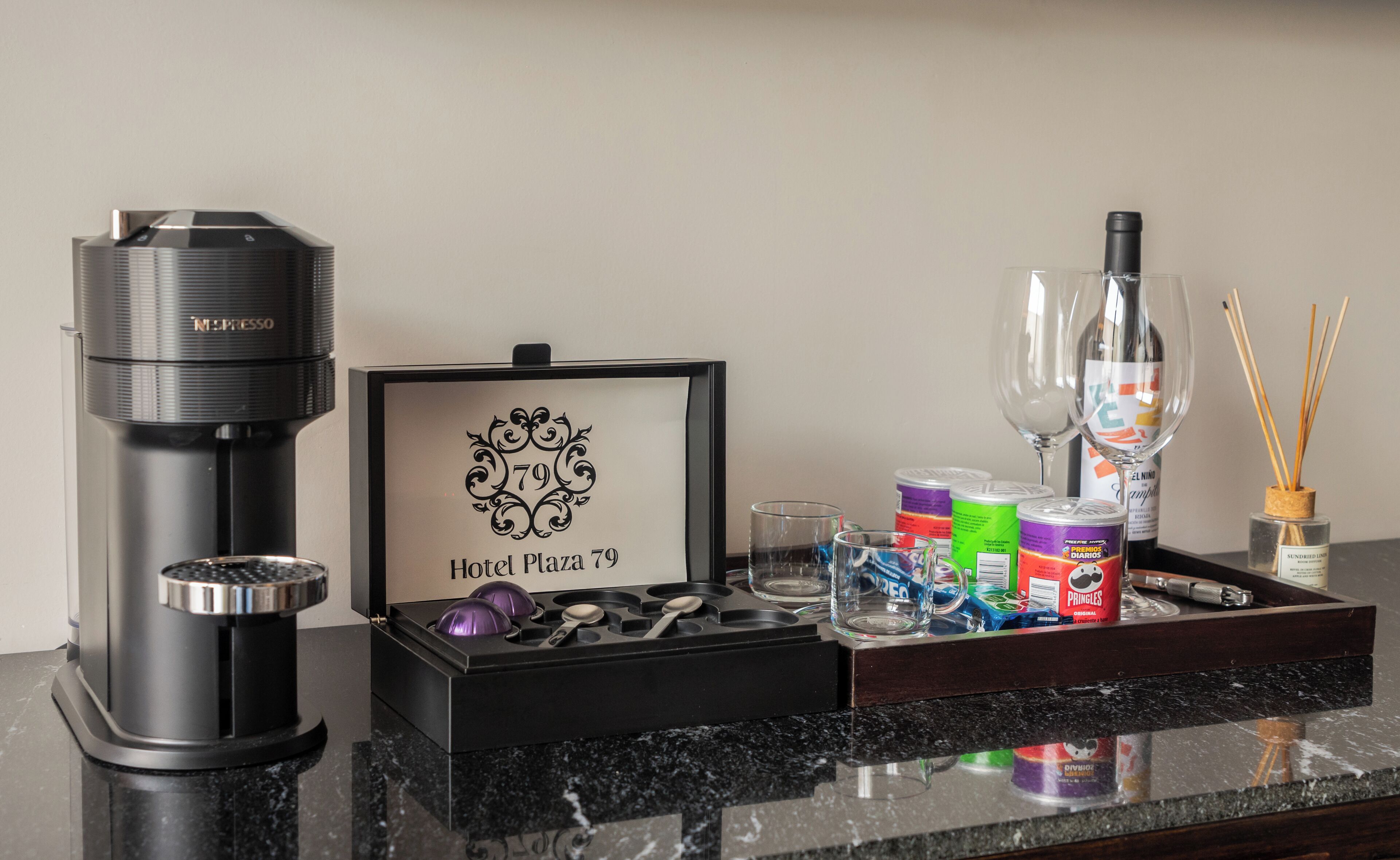 A minibar setup with a Nespresso machine, a framed hotel logo, a tray with snacks, glasses, a bottle, and a candle holder.