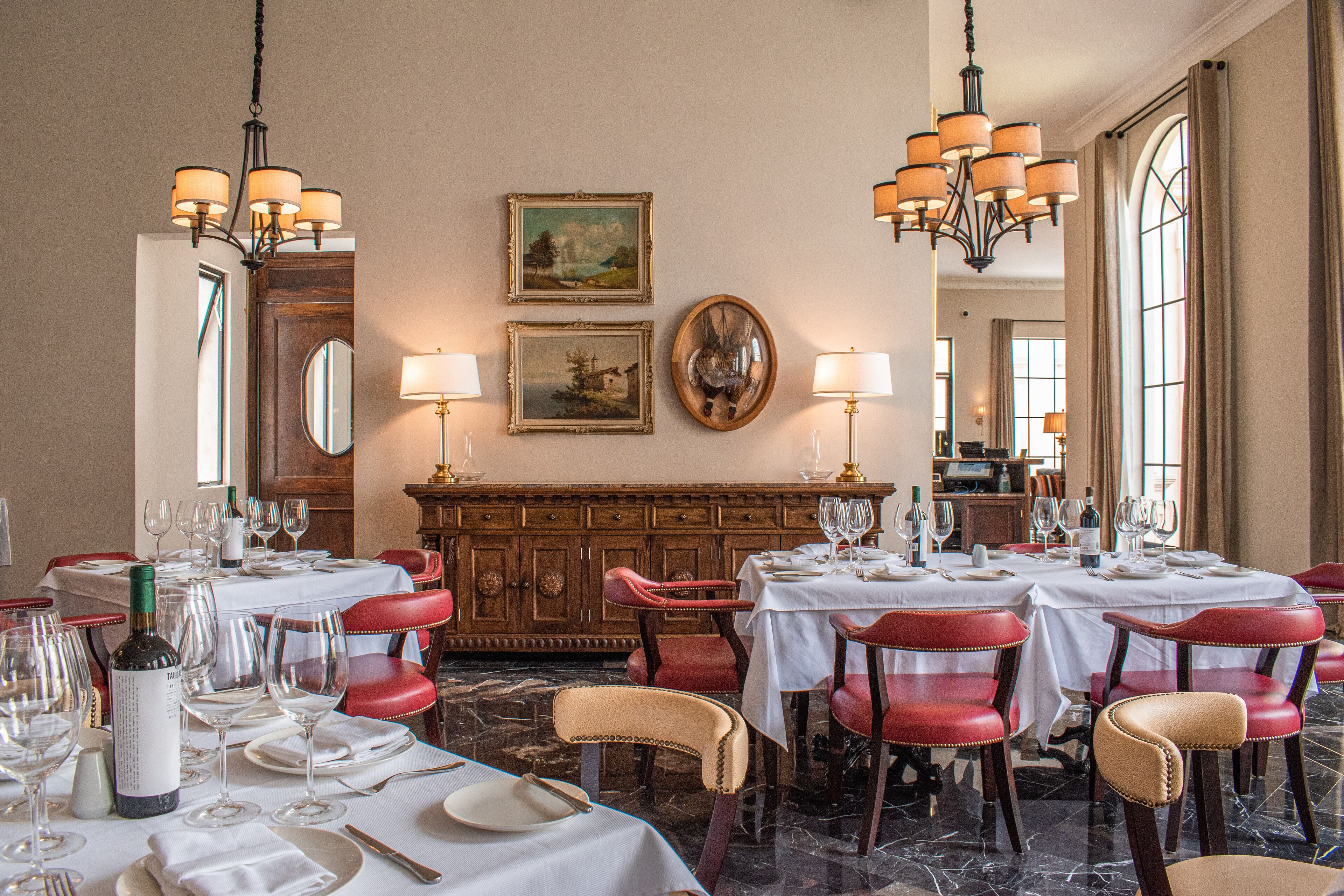 A dining area with tables set for a meal, chairs, chandeliers, and artwork on the walls.