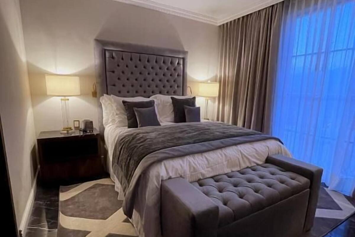 A modern bedroom with a tufted headboard, a large bed with a grey upholstered bench, a nightstand, and a floor-to-ceiling window with curtains.