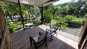 Ein Balkon mit Holzboden, Tisch und zwei Stühlen. Blick auf einen üppigen Garten mit verschiedenen Pflanzen und Bäumen.