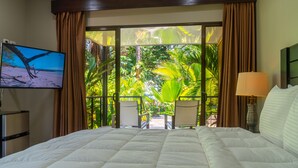 Ein Hotelzimmer mit einem großen Bett, einem Fernseher links, Blick auf üppiges Grün durch das Fenster und einer Lampe rechts.