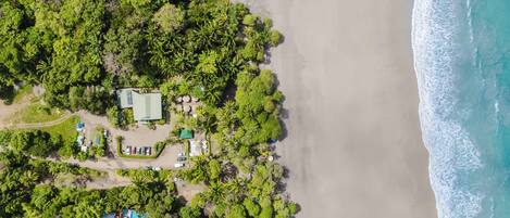 An aerial view of a coastal property with a beach, dense greenery, and several buildings.