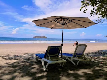 A beach with two lounge chairs under a large umbrella, a sandy beach, and the ocean in the background.