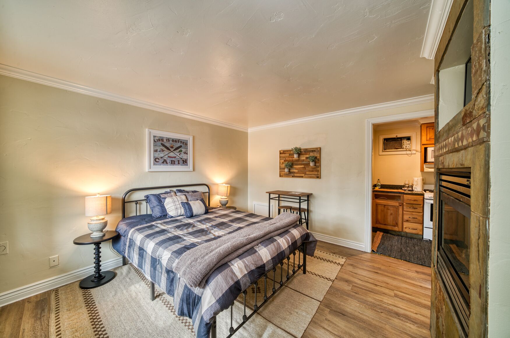 Een slaapkamer met een bed, nachtkastjes, een aan de muur hangend schilderij en een keukenruimte zichtbaar door een open deur.
