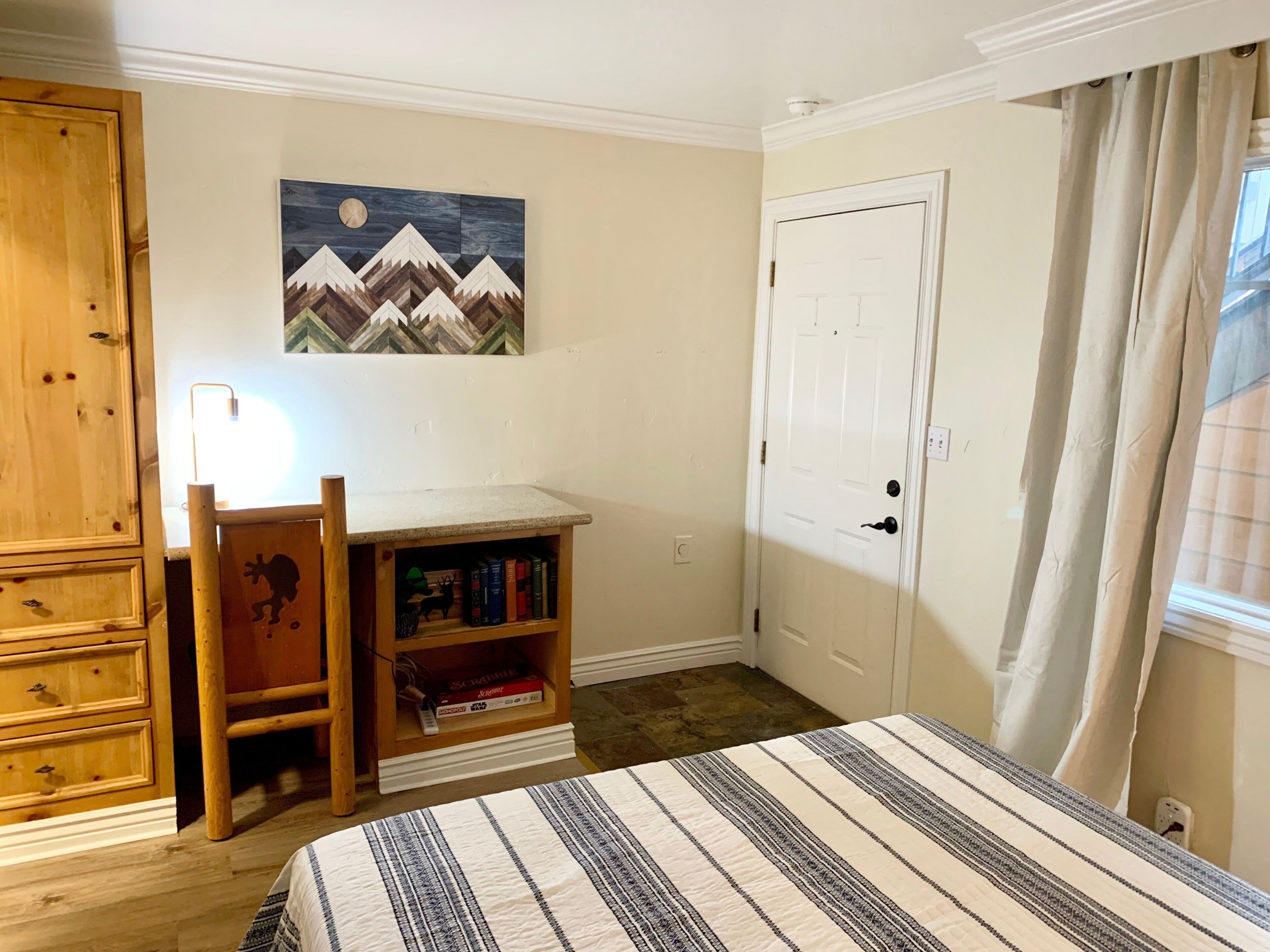 A bedroom with a bed, a desk with books, a wooden wardrobe, a painting, and a door.