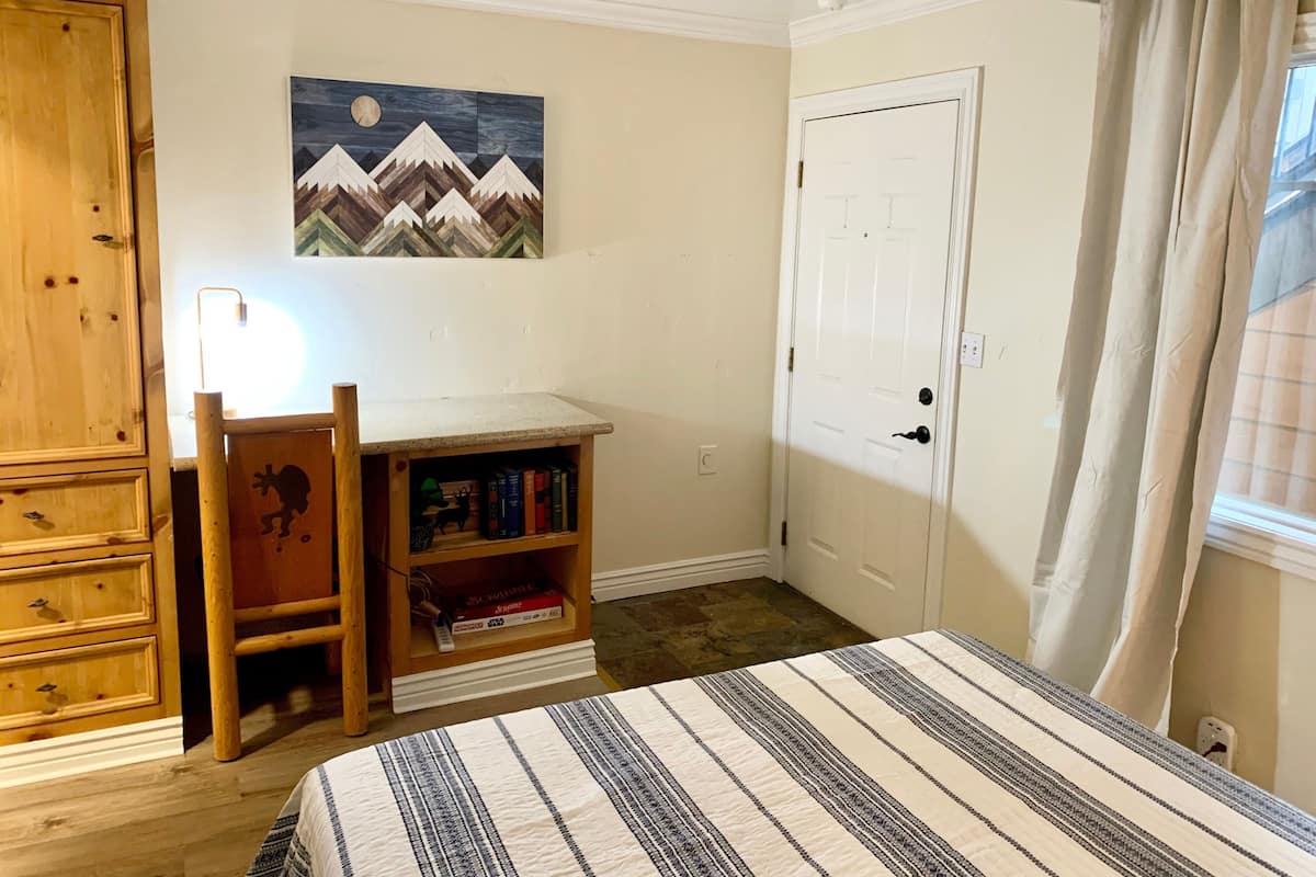 Um quarto com uma cama, uma escrivaninha com cadeira, uma estante de livros, uma porta e um quadro na parede.