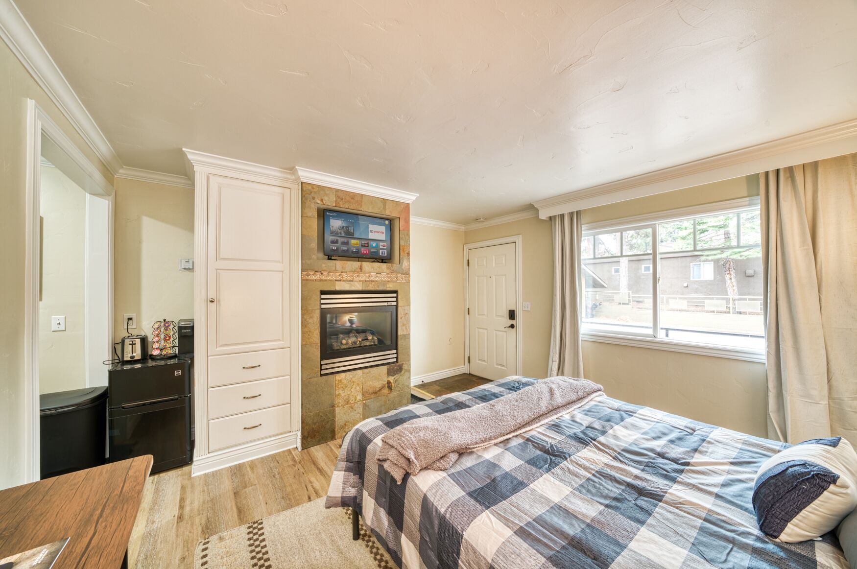 A bedroom with a bed, a television mounted above a fireplace, a desk, and a window with curtains.
