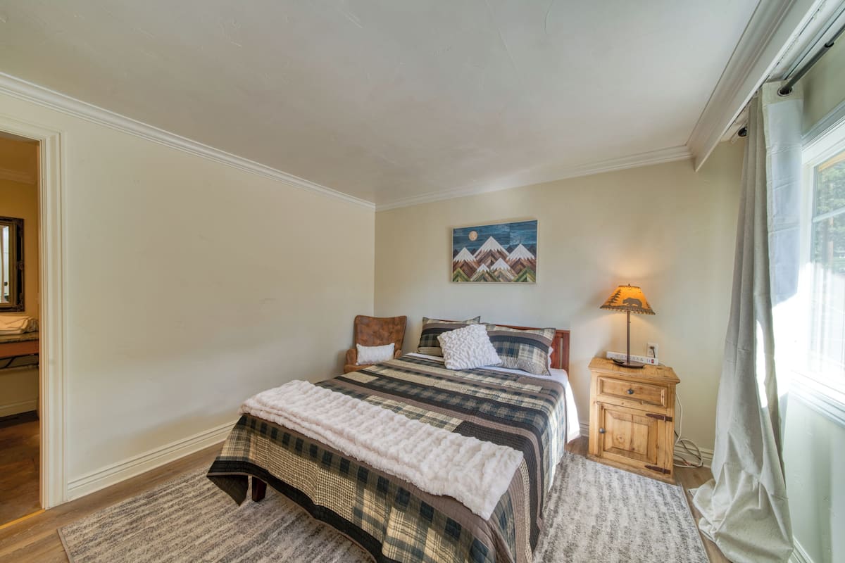 Quarto com cama, mesa de cabeceira, luminária, pintura e janela com cortinas.