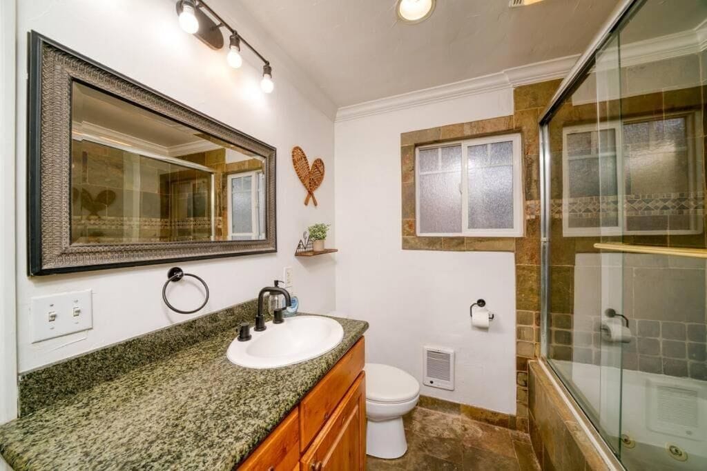 A bathroom with a granite countertop sink, a large mirror, a toilet, and a glass shower enclosure.