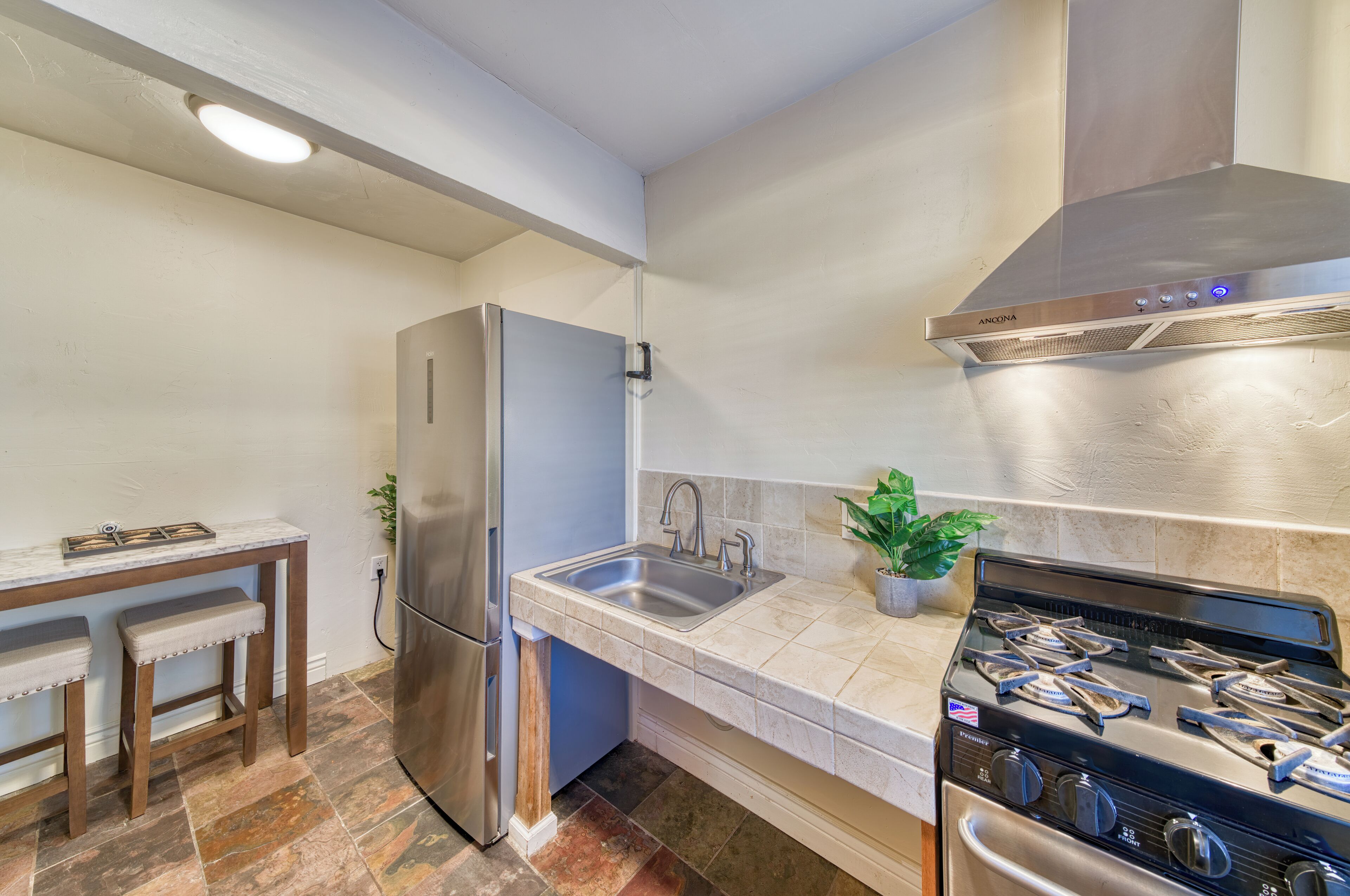 A compact kitchen with a stainless steel refrigerator, a sink, a stove, and a small dining area with stools.