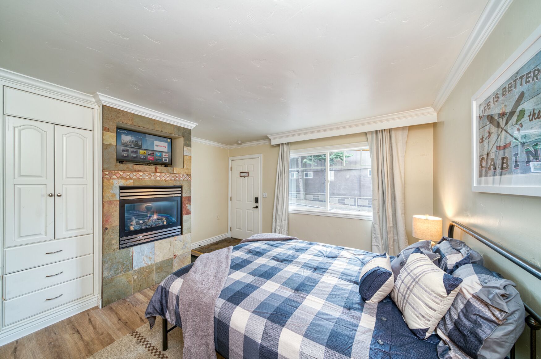 Ein Schlafzimmer mit Kamin, einem Bett mit kariertem Überwurf und einem Fernseher über dem Kamin. Ein Fenster mit Vorhängen ist ebenfalls zu sehen.