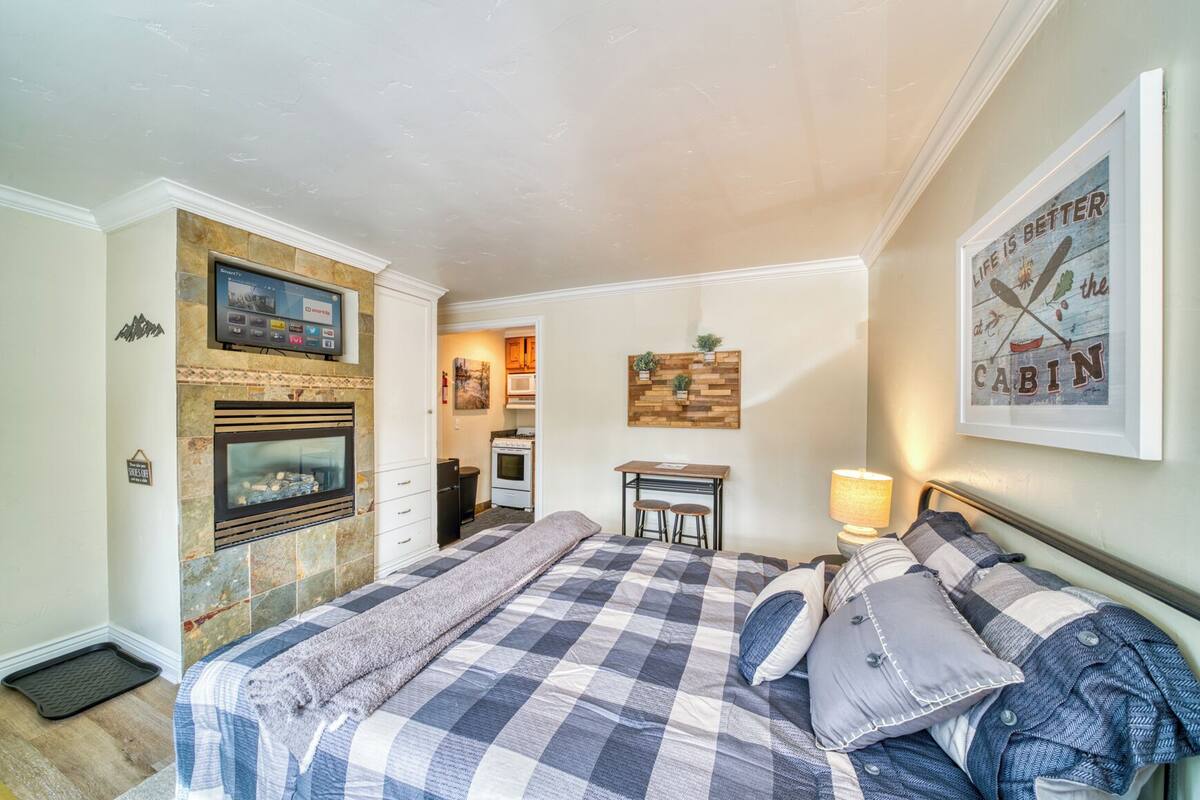 Um quarto aconchegante com lareira, cama com colcha xadrez, escrivaninha com duas cadeiras e uma TV fixada na parede.