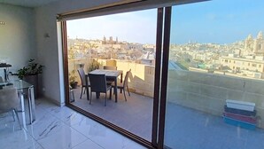Un balcon avec une table et des chaises, un pot de fleurs et une vue sur le paysage urbain, visible à travers de grandes portes vitrées.