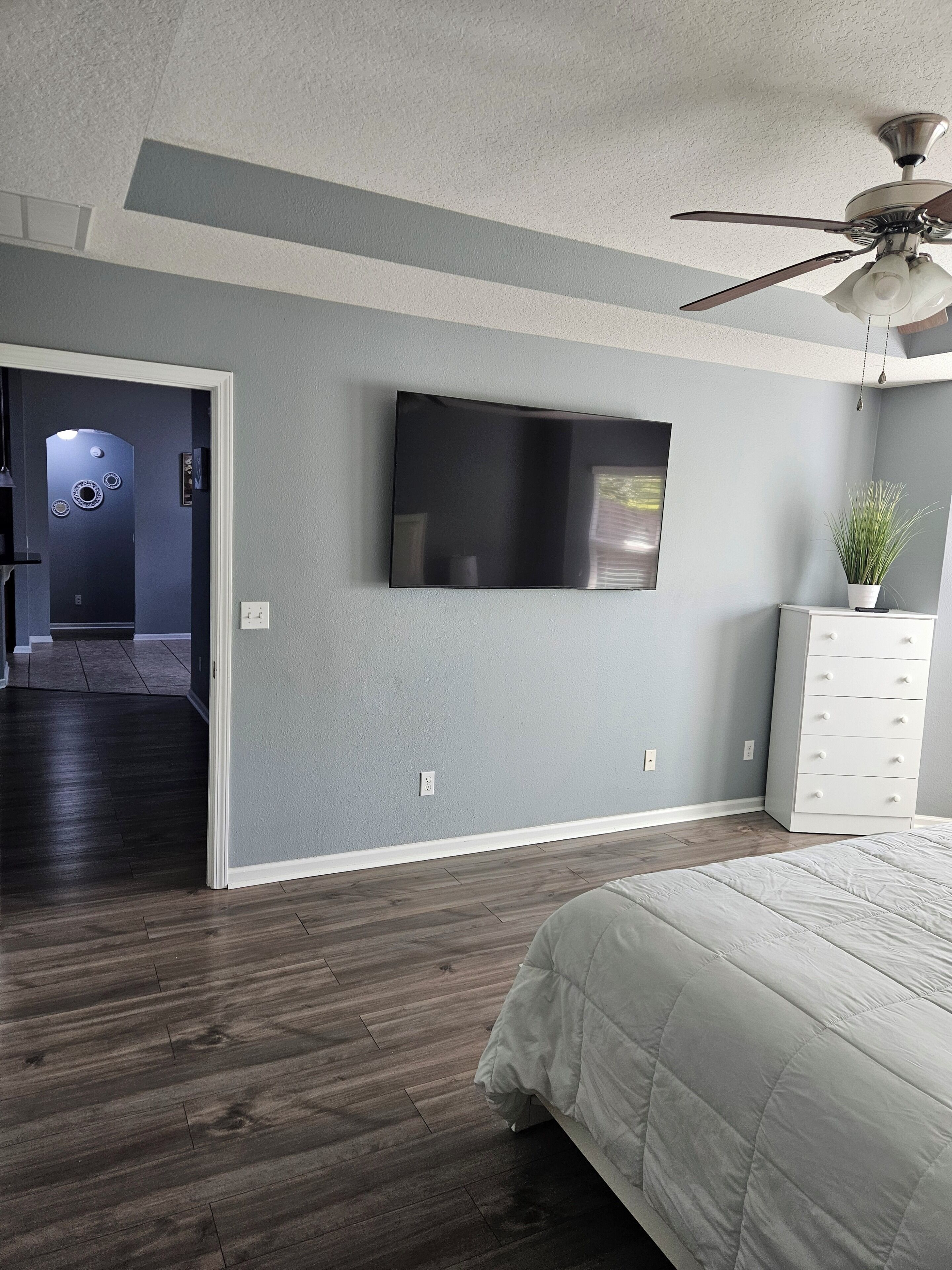 A bedroom with a bed, a dresser, a television on the wall, and a ceiling fan.