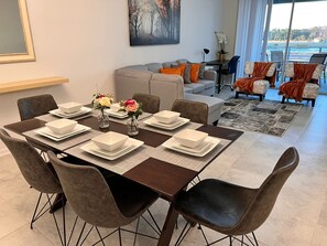 A dining area with a table set for four, surrounded by chairs. There is a sofa and armchairs in the background, and a large window with a view of the outdoors.