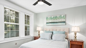 A bedroom with a large bed, two bedside tables with lamps, a ceiling fan, a window with blinds, and a painting on the wall.