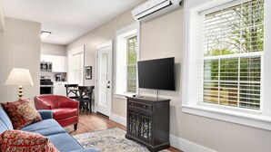 A living room with a blue sofa, red chair, and a TV on a stand. There is a kitchen in the background with white cabinets and a microwave.