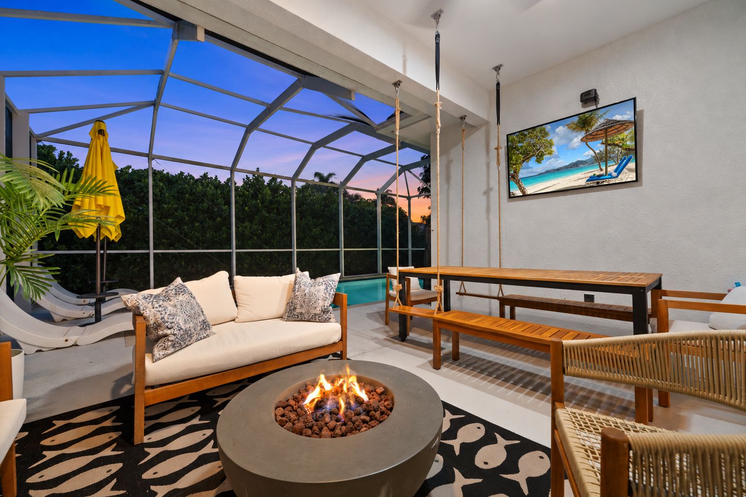 A modern outdoor living space with a sofa, wooden furniture, a fire pit, and a large window overlooking a pool.