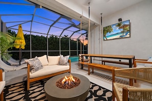 A modern outdoor living space with a sofa, wooden furniture, a fire pit, and a large window overlooking a pool.