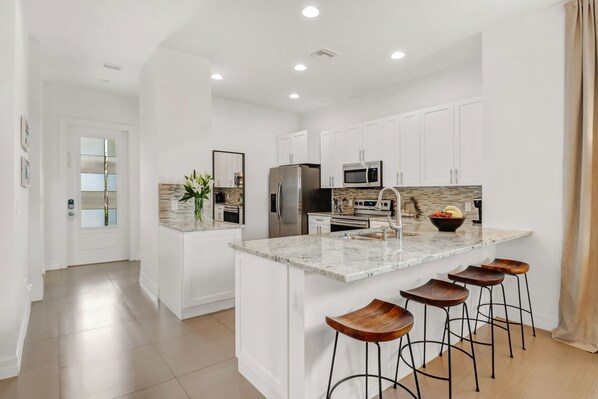 Uma cozinha moderna com armários brancos, bancada de granito, eletrodomésticos de aço inoxidável e uma ilha central com banquetas.
