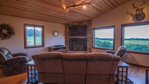 Un salon chaleureux avec une cheminée, des fauteuils en cuir inclinables, une tête de cerf accrochée au mur et une vue sur un paysage verdoyant.