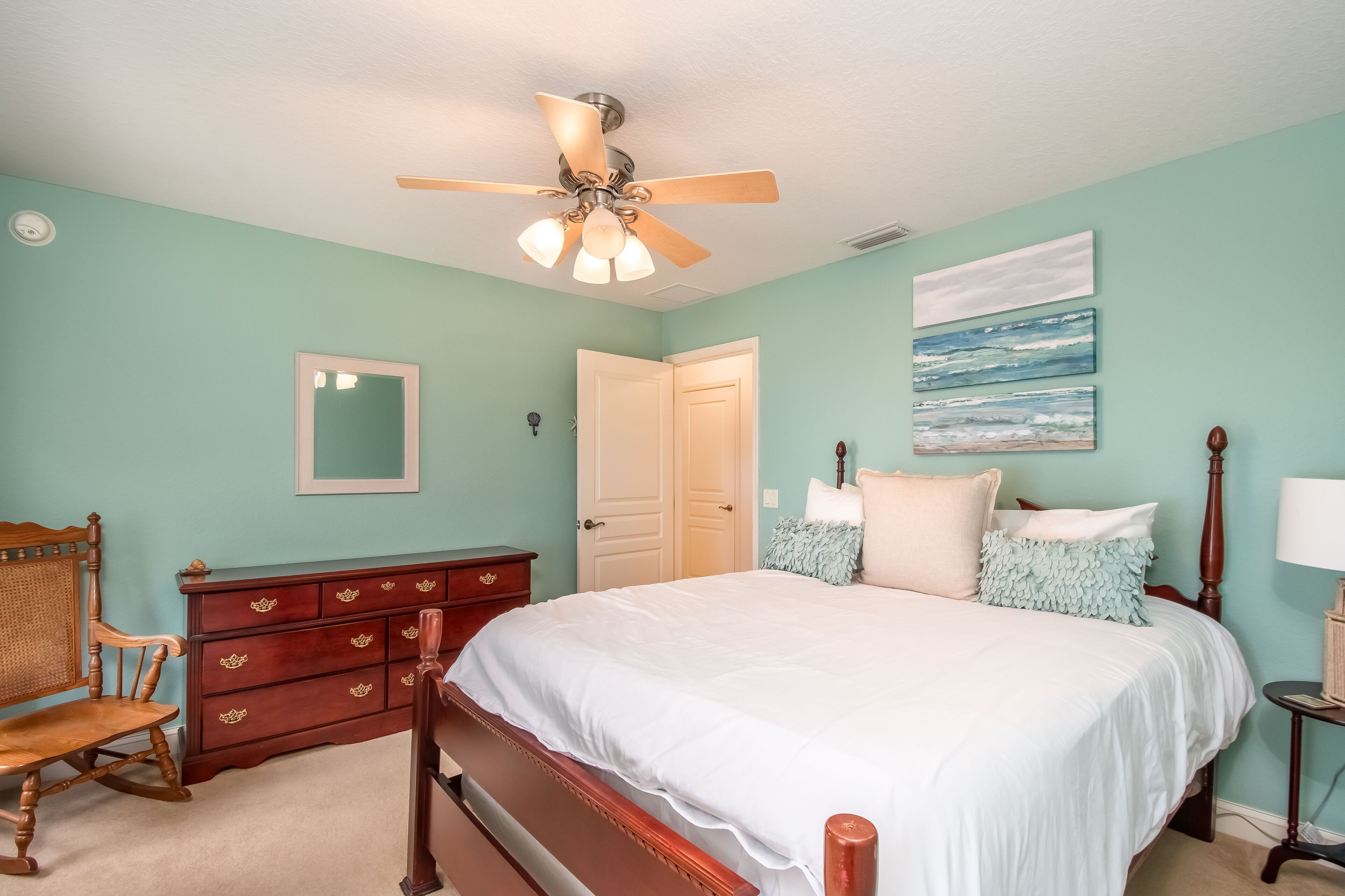 A bedroom with a bed, wooden dresser, chair, ceiling fan, and artwork on the wall.