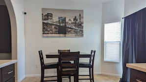 Een eethoek met een tafel en stoelen, een stadsgezichtschilderij aan de wand, een raam met jaloezieën en een donker kastje.