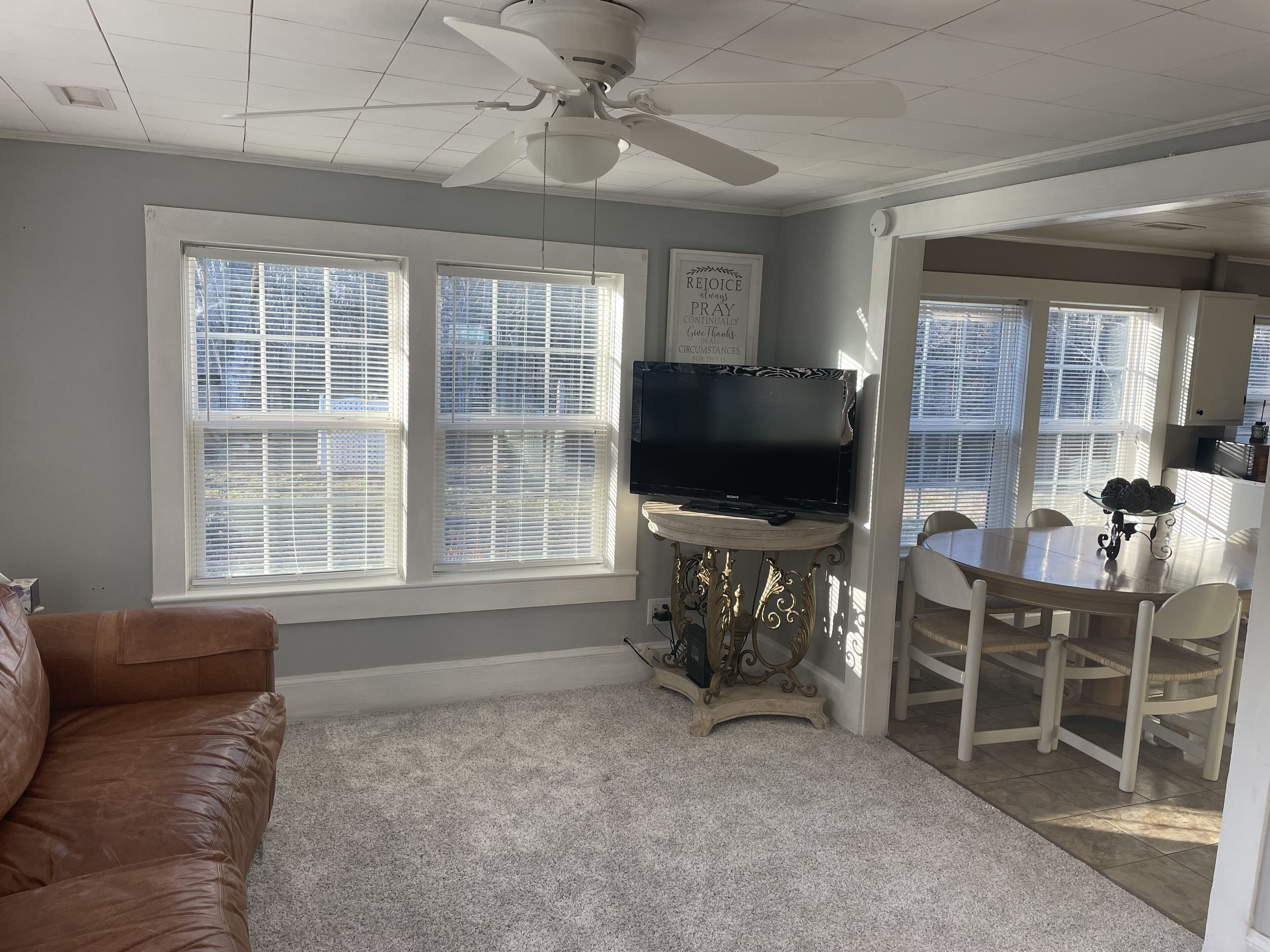 A living room with a brown leather couch, a TV on a stand, a dining table with chairs, and a ceiling fan.