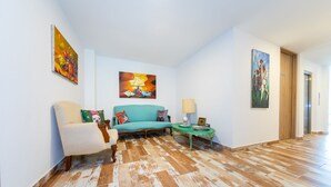 Una habitación con un sofá verde azulado, un sillón color beige y suelo de madera. Hay cuadros en las paredes y una lámpara sobre una mesita.