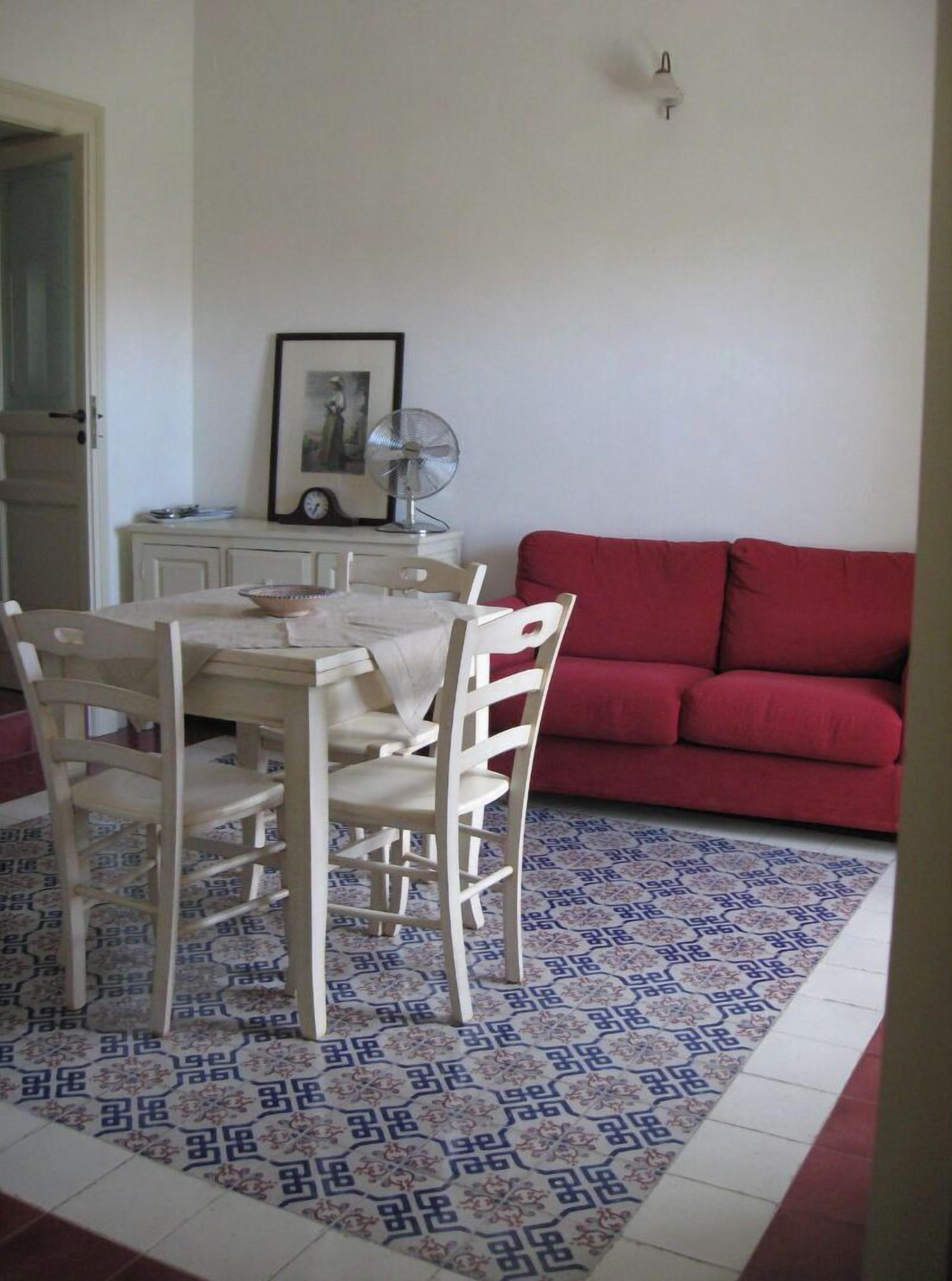 A dining area with a table and chairs, a red sofa, a patterned rug, and a wall-mounted fan.