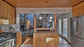 Une cuisine moderne avec des meubles en bois, des appareils en acier inoxydable, des luminaires suspendus et une vue sur l’espace de vie.
