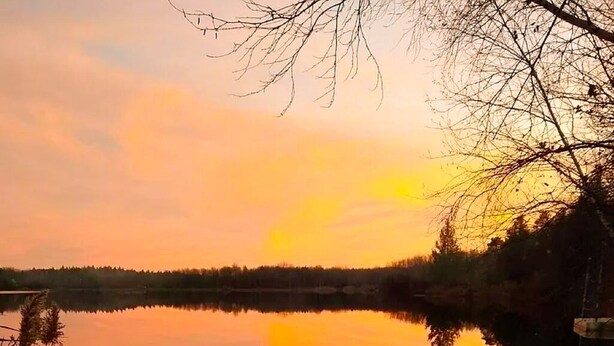 Une terrasse en bois avec un foyer et un verre d’eau, donnant sur un lac paisible au coucher du soleil.