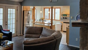 Un vaste espace de vie doté d’un haut plafond, de poutres apparentes, d’un ventilateur de plafond et offrant une vue sur la cuisine grâce à une porte ouverte.