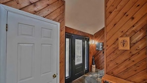 Un couloir intérieur aux murs en bois, avec une porte blanche, une porte noire dotée de panneaux de verre et un luminaire fixé au mur.
