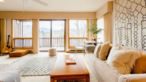 A spacious living room with a large sofa, a coffee table, and a guitar. There are large windows with a view of the outdoors.