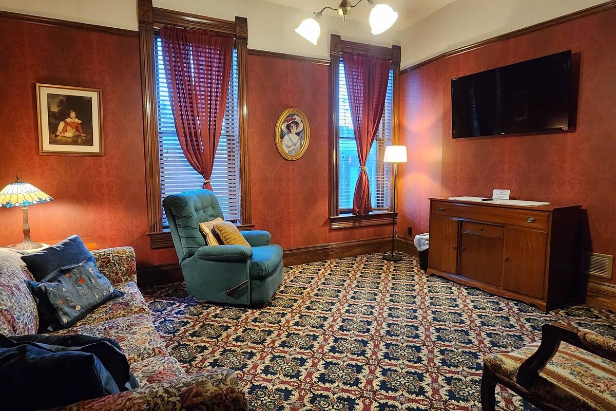 A room with a floral-patterned sofa, a green armchair, a patterned carpet, a wooden cabinet, and a television mounted on the wall.