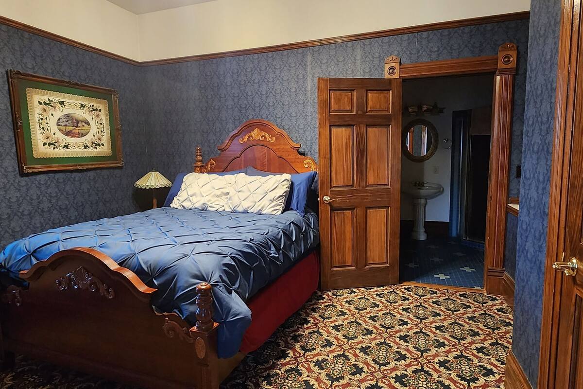 A bedroom with a wooden bed, a framed picture, a window with blinds, and a patterned carpet.