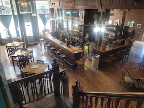 A bar area with wooden tables and chairs, a counter with various bottles, and a staircase leading to another level.