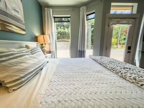 Quarto com cama, mesa de cabeceira, luminária e vista do exterior através de uma porta.