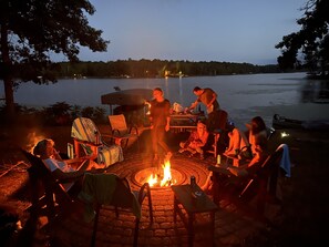 A group of people around a campfire with a lake in the background.