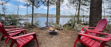 A lakeside setting with red Adirondack chairs arranged around a campfire.