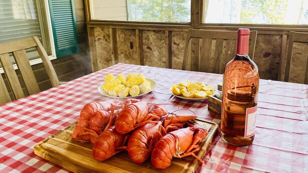 Uma mesa com toalha xadrez, tábua de madeira com lagostas, espigas de milho e fatias de limão, e uma garrafa de vinho rosa.