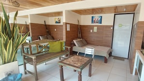 Ein gemütliches Zimmer mit einer Holzbank, einem kleinen Tisch und einer Couch. An den Wänden hängen Gemälde und eine Tür ist mit „02 Quarto“ beschriftet.